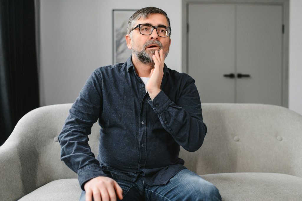 Elderly Man Having Toothache Touching Cheek Suffering From Pain Sitting On Sofa