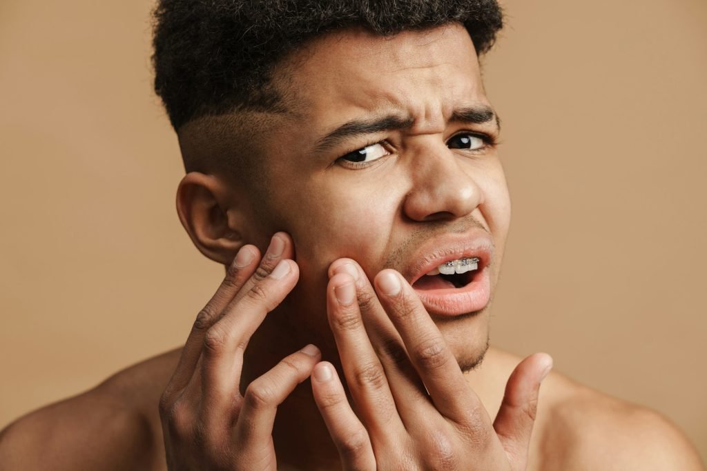 Young shirtless man frowning while squeezing pimple on his face